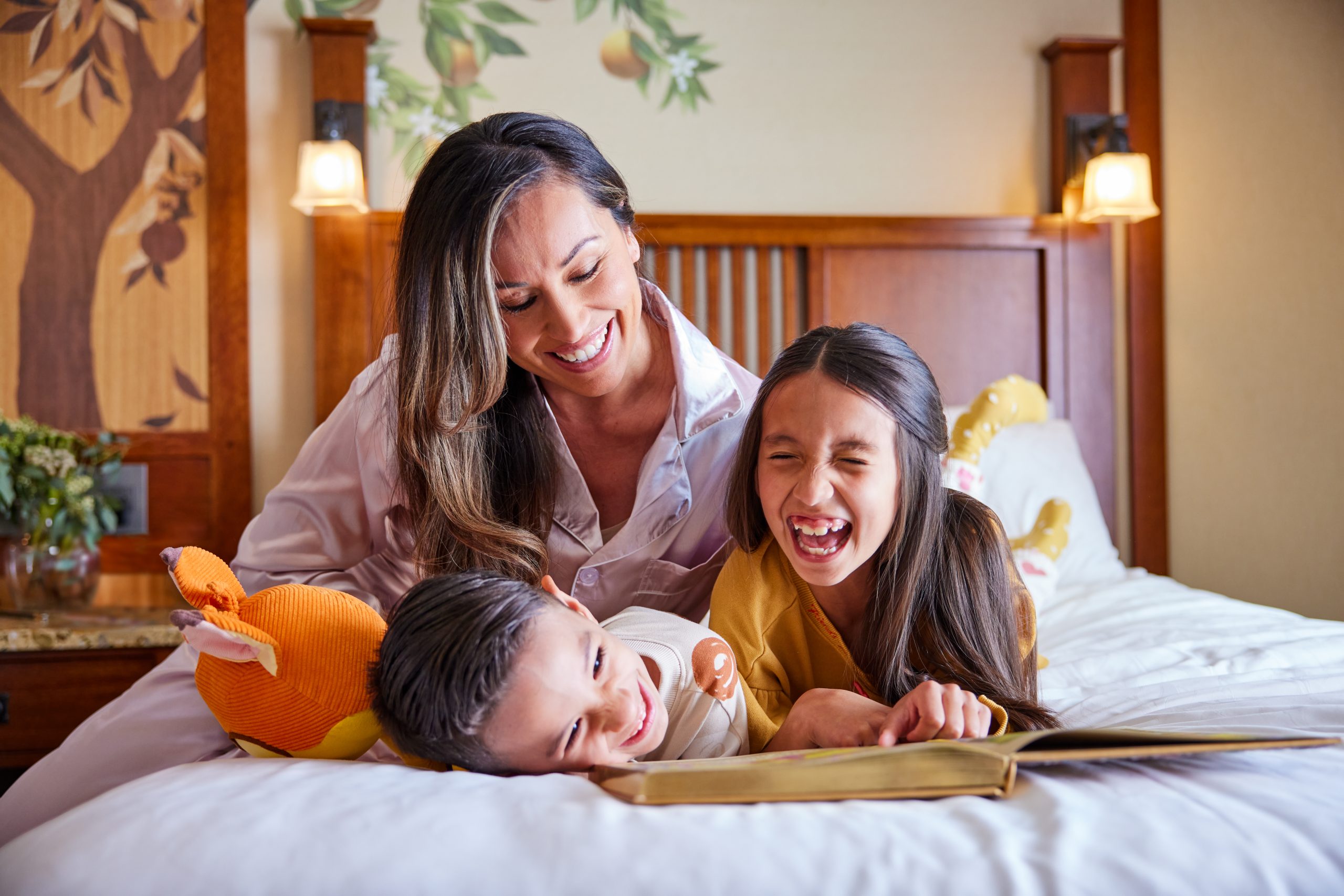 A woman and two small children read a book together on the hotel bed and laugh. 